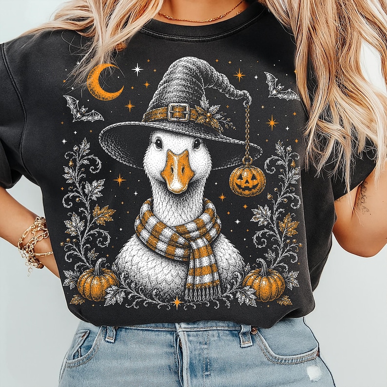 May include: A black crewneck sweatshirt featuring a graphic illustration of a white goose wearing a pointed witch hat and a striped orange and white scarf. The design includes a hanging jack-o'-lantern, bats, a crescent moon, and pumpkins.