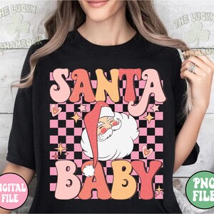May include: Black t-shirt with a retro design featuring the words "SANTA BABY" in pink, orange, and peach. Santa Claus is in the center, with a pink and black checkered background, hearts, and stars.