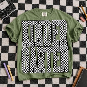 K&ouml;nnte beinhalten: Ein olivgr&uuml;nes Kurzarm-T-Shirt mit dem Wort KINDERGARTEN, das in gro&szlig;en, fetten, schwarz-wei&szlig; karierten Blockbuchstaben in der Mitte aufgedruckt ist. Das T-Shirt hat einen Rundhalsausschnitt und eine Standardpassform mit einem sichtbaren Markenetikett.