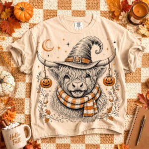 May include: A beige t-shirt featuring a black and white line art illustration of a Highland cow wearing a witch hat and a striped scarf. The cow has small jack-o'-lanterns hanging from its horns, surrounded by stars, a crescent moon, and leaves.