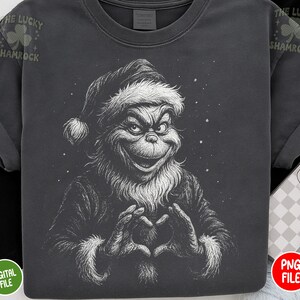 May include: Dark gray t-shirt featuring a detailed illustration of a Grinch-like character forming a heart with their hands. The character is wearing a Santa hat and coat with white trim. The text "THE LUCKY SHAMROCK" is printed on the top left.
