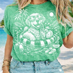May include: Green t-shirt featuring a white graphic of a dog wearing sunglasses and floating in a pool on an inner tube. The dog is holding a drink with a straw. Palm trees, a sun, and a beach ball are also in the design.