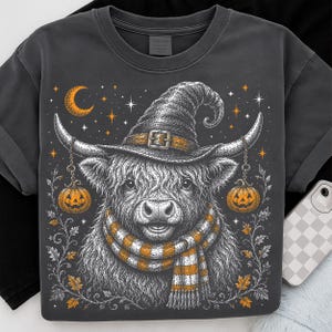 May include: A charcoal gray t-shirt featuring a detailed graphic of a Highland cow wearing a witch hat and a striped scarf. The cow is flanked by two hanging jack-o'-lanterns, with a crescent moon and stars in the background, all rendered in orange and white.