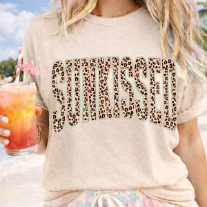 May include: A beige short-sleeved t-shirt featuring the word SUNKISSED printed in a bold, arched font. The letters have a brown and black leopard print pattern with a thin teal outline. The shirt has a crew neckline and a relaxed, casual fit.