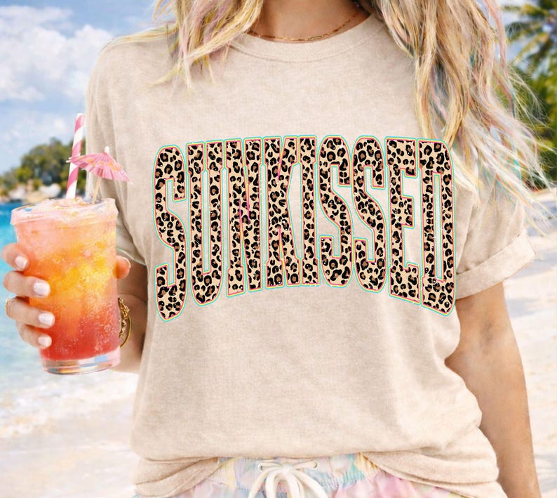 May include: A beige short-sleeved t-shirt featuring the word SUNKISSED printed in a bold, arched font. The letters have a brown and black leopard print pattern with a thin teal outline. The shirt has a crew neckline and a relaxed, casual fit.