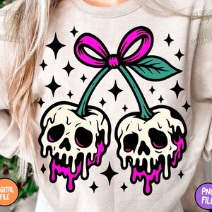 May include: A beige sweatshirt featuring a graphic of two cherries designed as skulls with pink dripping accents. The cherries are connected by green stems and a pink bow. Black star shapes are scattered around the design.