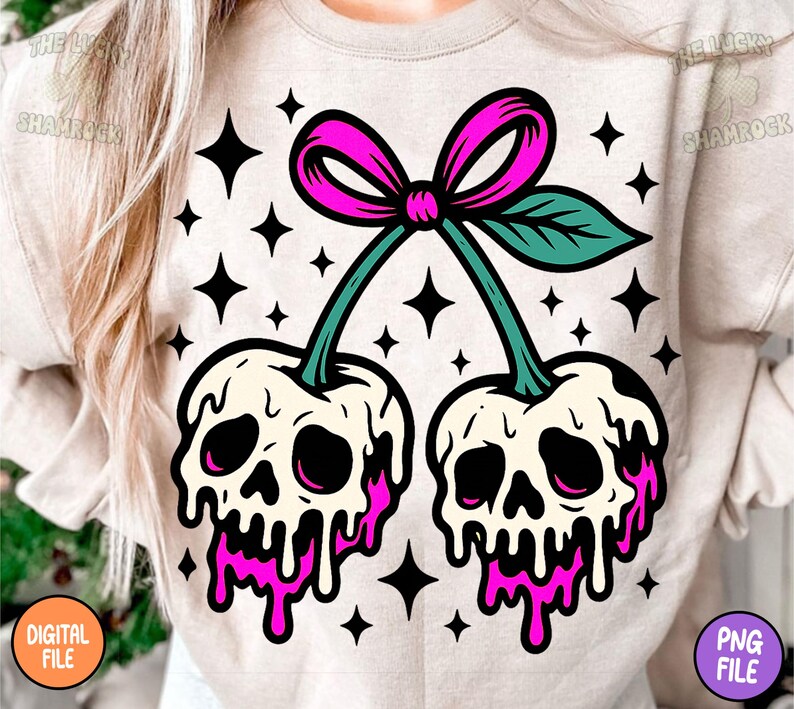 May include: A beige sweatshirt featuring a graphic of two cherries designed as skulls with pink dripping accents. The cherries are connected by green stems and a pink bow. Black star shapes are scattered around the design.