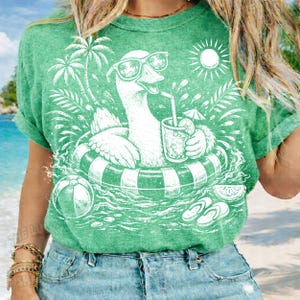 May include: Green t-shirt featuring a white graphic of a goose wearing sunglasses and floating in a pool on an inner tube. The goose is holding a drink with a straw. Palm trees, a sun, and a beach ball are also in the design.
