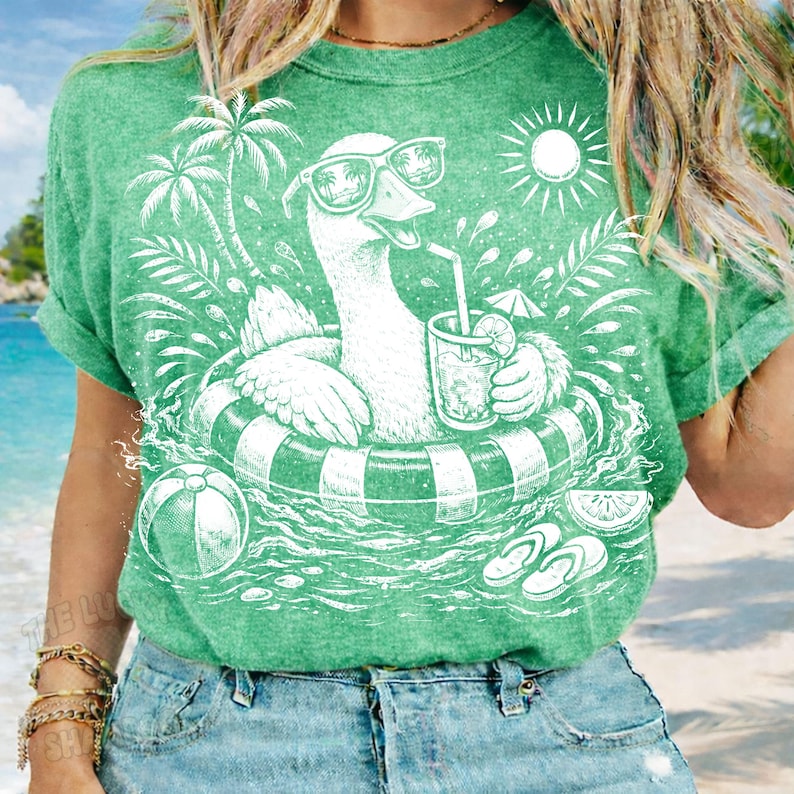 May include: Green t-shirt featuring a white graphic of a goose wearing sunglasses and floating in a pool on an inner tube. The goose is holding a drink with a straw. Palm trees, a sun, and a beach ball are also in the design.