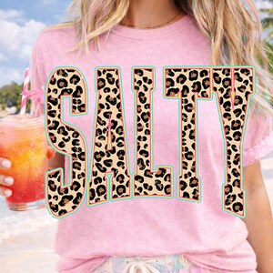 May include: Pink t-shirt with the word "SALTY" in large, leopard print letters outlined in turquoise. The person is holding a tropical drink with a pink straw and a small umbrella.