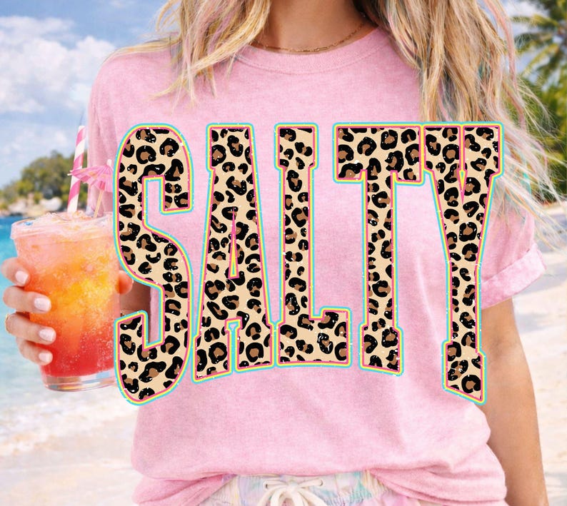May include: Pink t-shirt with the word "SALTY" in large, leopard print letters outlined in turquoise. The person is holding a tropical drink with a pink straw and a small umbrella.