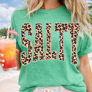 May include: A green t-shirt with the word "SALT" in large, leopard print letters. The person is holding a tropical drink with a pink umbrella and a straw. The shirt has rolled-up sleeves.