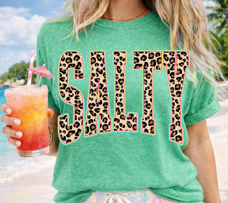 May include: A green t-shirt with the word "SALT" in large, leopard print letters. The person is holding a tropical drink with a pink umbrella and a straw. The shirt has rolled-up sleeves.