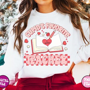 May include: White sweatshirt with a pink and red graphic design that says "Cupid's Favorite Teacher" with a heart, a book, and a pencil.