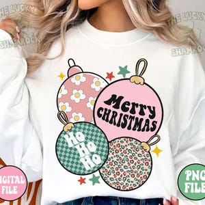 May include: A white sweatshirt featuring a graphic of four Christmas ornaments in pink, green, and leopard print. The ornaments have the text "Merry Christmas" and "Ho Ho Ho". The design includes stars and daisy patterns.