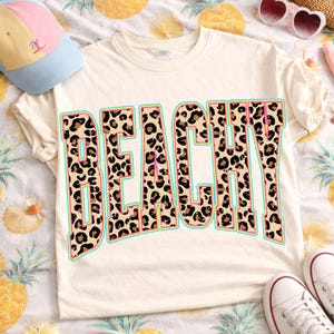 May include: Cream-colored t-shirt with the word "BEACHY" in a leopard print design with a teal outline and pink accents. A light blue and pink baseball cap is visible in the upper left corner.