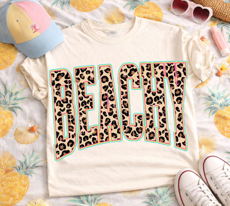 May include: Cream-colored t-shirt with the word "BEACHY" in a leopard print design with a teal outline and pink accents. A light blue and pink baseball cap is visible in the upper left corner.