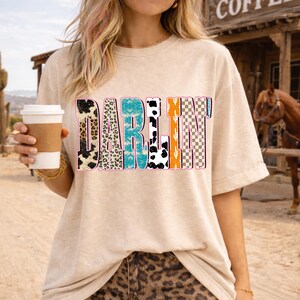 May include: A beige t-shirt with the word "DARLIN" in a colorful, patterned design. The letters feature animal prints, turquoise, orange, and checkered patterns. The person is holding a coffee cup.