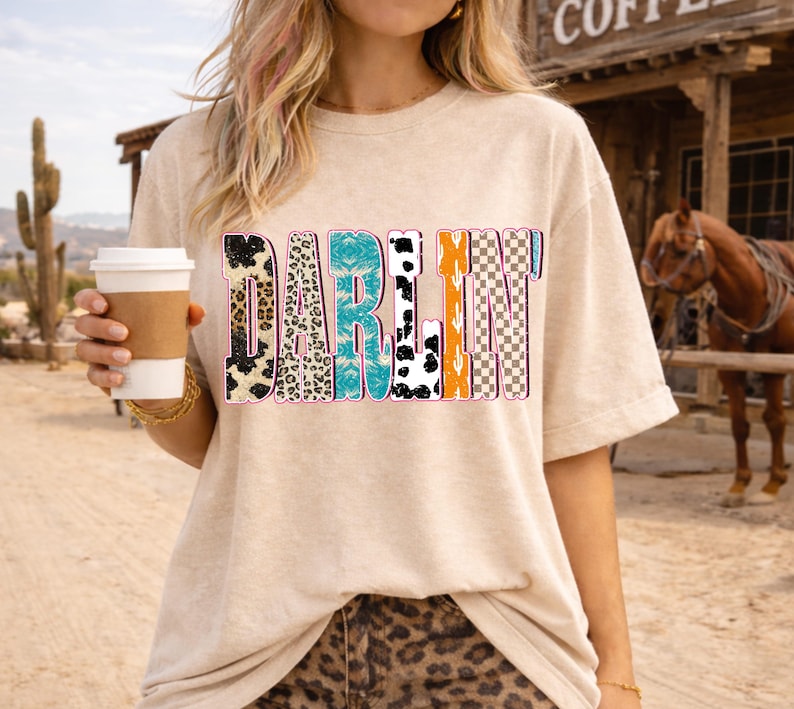May include: A beige t-shirt with the word "DARLIN" in a colorful, patterned design. The letters feature animal prints, turquoise, orange, and checkered patterns. The person is holding a coffee cup.