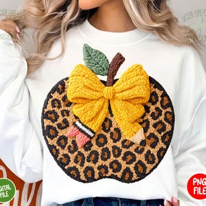 May include: White sweatshirt featuring a large, textured apple design. The apple has a leopard print pattern, a yellow bow, a green leaf, and a brown stem. A pencil and eraser are also included in the design. The words "THE LUCKY SHAMROCK" are printed on the top.