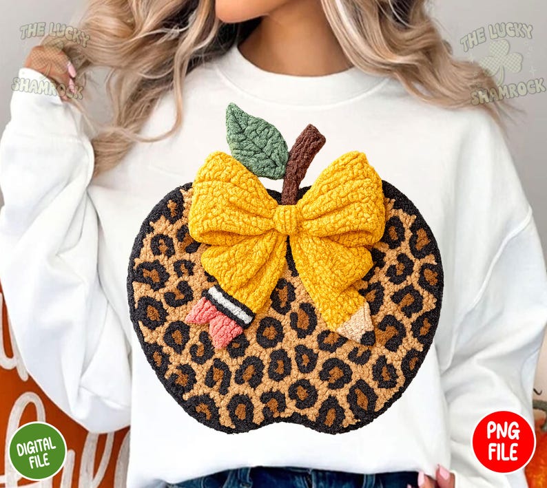 May include: White sweatshirt featuring a large, textured apple design. The apple has a leopard print pattern, a yellow bow, a green leaf, and a brown stem. A pencil and eraser are also included in the design. The words "THE LUCKY SHAMROCK" are printed on the top.