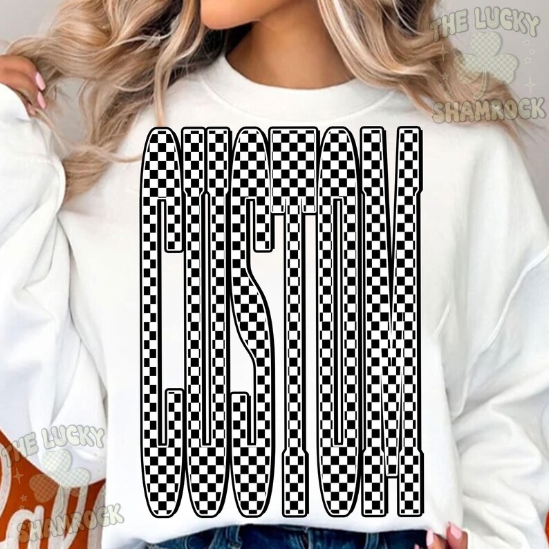 May include: White sweatshirt featuring the word "CUSTOM" in a black and white chequered pattern. The design is in a vertical, block letter style. The sweatshirt has a crew neck and long sleeves.