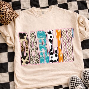 May include: Cream-colored t-shirt with the word "DARLIN" in a colorful, patterned design. The patterns include animal prints, turquoise, orange, and checkered designs. A leopard print hat and checkered sneakers are also visible.