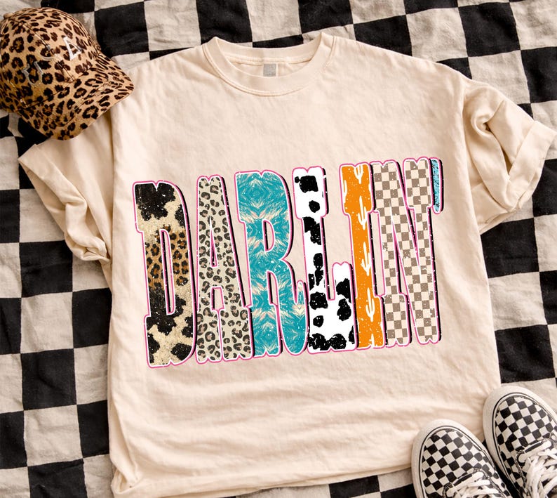 May include: Cream-colored t-shirt with the word "DARLIN" in a colorful, patterned design. The patterns include animal prints, turquoise, orange, and checkered designs. A leopard print hat and checkered sneakers are also visible.