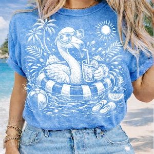 May include: Blue t-shirt featuring a white graphic of a flamingo wearing sunglasses and floating in a pool with a drink. The design includes palm trees, a beach ball, and the sun, evoking a summer theme.