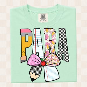 K&ouml;nnte beinhalten: Ein helles mintgr&uuml;nes T-Shirt mit dem Wort PARA in gro&szlig;en Blockbuchstaben. Jeder Buchstabe hat ein einzigartiges Muster: Bleistift, liniertes Papier, Blumenmuster und schwarz-wei&szlig;es Schachbrettmuster. Darunter eine gro&szlig;e Schleife mit rotem Apfel.