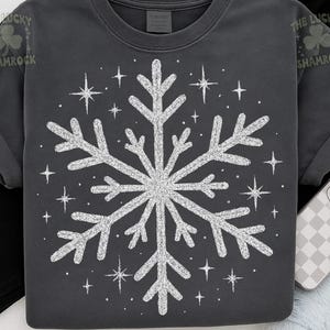 May include: Dark gray t-shirt featuring a large, glittery white snowflake design. The snowflake is surrounded by smaller starbursts. The Lucky Shamrock logo is visible on the upper left and right of the shirt. This winter-themed shirt is perfect for the holiday season.