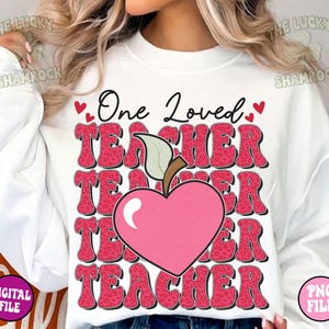 May include: White sweatshirt with a pink and red graphic design that says "One Loved Teacher" with a pink heart and apple design.