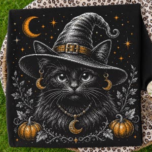 May include: A black t-shirt featuring a graphic illustration of a black cat wearing a pointed witch hat with a buckle, gold crescent moon earrings, and a gold crescent moon necklace, surrounded by orange pumpkins, stars, and white leaf outlines.