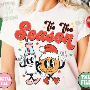May include: White t-shirt with a retro design featuring the text "Tis The Season" in red and brown. The graphic includes a cartoon milk bottle and cookie, both wearing Santa hats and giving peace signs. Stars and shamrocks are also included.