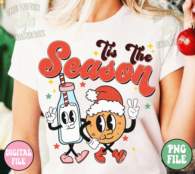 May include: White t-shirt with a retro design featuring the text "Tis The Season" in red and brown. The graphic includes a cartoon milk bottle and cookie, both wearing Santa hats and giving peace signs. Stars and shamrocks are also included.