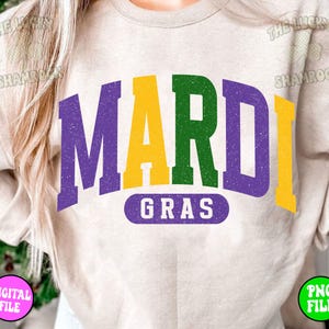 Puede incluir: Una sudadera beige claro con el texto "Mardi Gras" en letras moradas, amarillas y verdes. Las letras est&aacute;n en una fuente de estilo vintage con un aspecto desgastado.