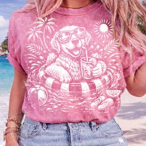 May include: Pink t-shirt featuring a white graphic of a golden retriever wearing sunglasses and relaxing in a pool float with a drink. The design includes palm trees, a beach ball, and the sun, evoking a summer theme.