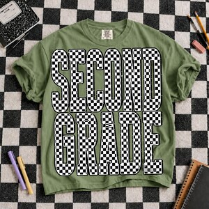 K&ouml;nnte beinhalten: Ein olivgr&uuml;nes Kurzarm-T-Shirt mit dem Text "SECOND GRADE", der in einem gro&szlig;en, schwarz-wei&szlig; karierten Muster gedruckt ist. Die Buchstaben haben ein abgenutztes, strukturiertes Aussehen und das T-Shirt hat einen Rundhalsausschnitt.