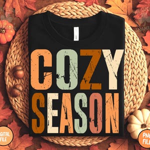 May include: Black t-shirt with the words "COZY SEASON" in large, distressed, orange, green, and cream-colored letters. The shirt is displayed on a woven mat surrounded by fall leaves, pumpkins, and acorns.