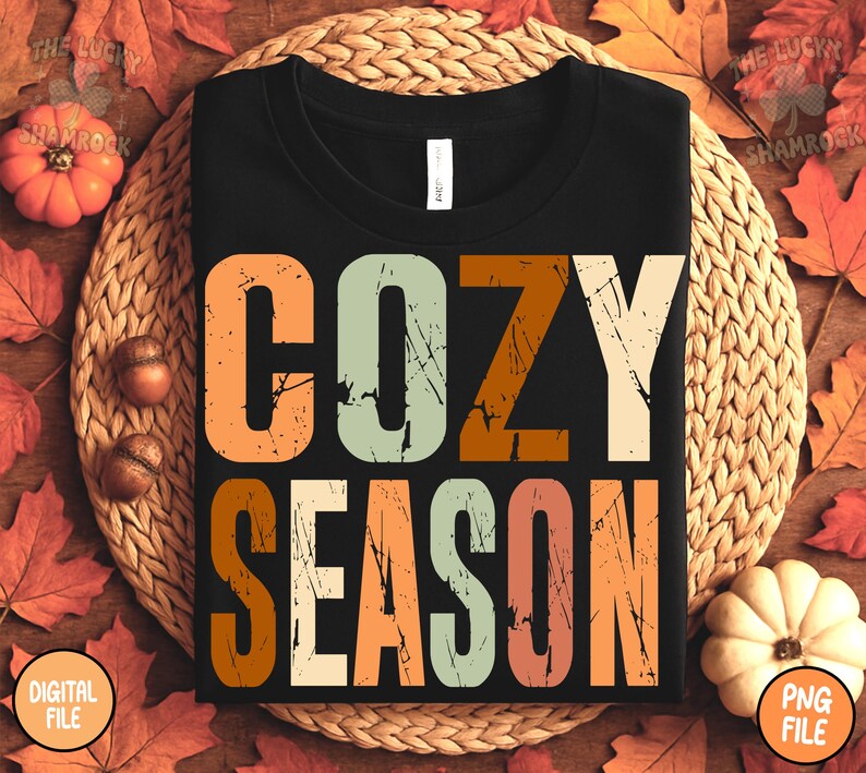 May include: Black t-shirt with the words "COZY SEASON" in large, distressed, orange, green, and cream-colored letters. The shirt is displayed on a woven mat surrounded by fall leaves, pumpkins, and acorns.