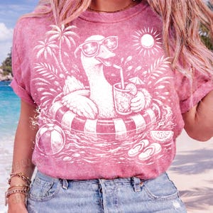 May include: Pink t-shirt featuring a white graphic of a goose wearing sunglasses and floating in a pool with a drink. The design includes a beach ball, palm trees, and a sun.