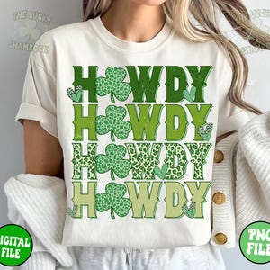 May include: A white t-shirt with the word "HOWDY" repeated four times in green, yellow, and brown. Each "HOWDY" is decorated with shamrock-shaped leaves. The shirt is being worn by a person.