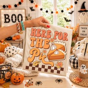 May include: A beige t-shirt featuring a graphic print with the text "HERE FOR THE PIE" in a retro orange font. The design includes two slices of pumpkin pie topped with whipped cream, colorful autumn leaves, and a brown and beige checkerboard pattern.