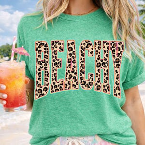 May include: A teal t-shirt with the word "BEACHY" in large, leopard print letters. The person is holding a tropical drink with a pink umbrella and a straw. The background shows a beach scene.