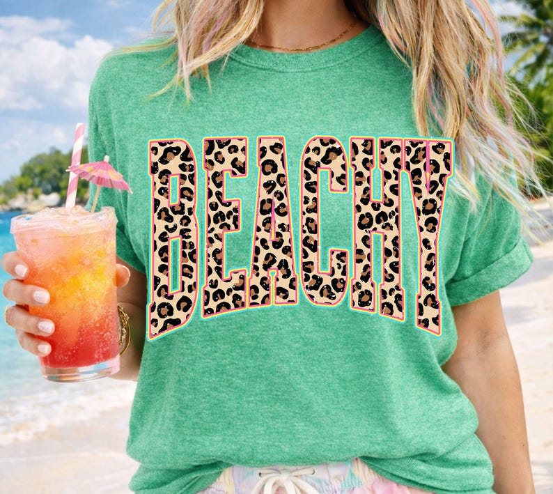 May include: A teal t-shirt with the word "BEACHY" in large, leopard print letters. The person is holding a tropical drink with a pink umbrella and a straw. The background shows a beach scene.