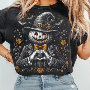 May include: A black t-shirt featuring a graphic illustration of a pumpkin-headed figure wearing a witch hat and a suit with a bow tie. The figure is making a heart shape with its hands, surrounded by orange maple leaves, bats, and stars.