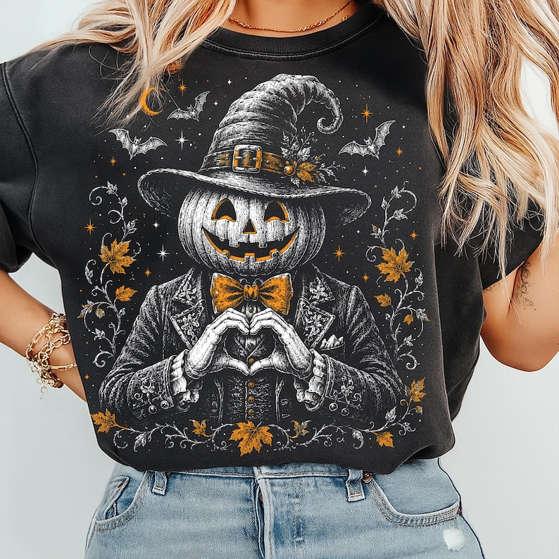 May include: A black t-shirt featuring a graphic illustration of a pumpkin-headed figure wearing a witch hat and a suit with a bow tie. The figure is making a heart shape with its hands, surrounded by orange maple leaves, bats, and stars.