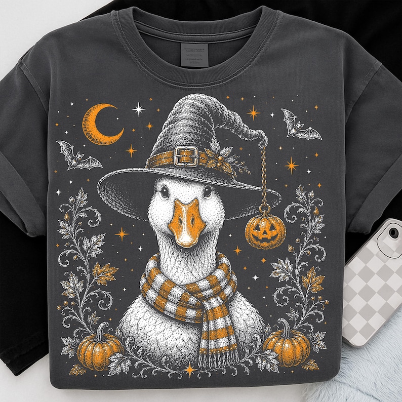 May include: A charcoal gray t-shirt featuring a graphic illustration of a white goose wearing a witch hat and a striped orange and white scarf. The design includes orange pumpkins, a crescent moon, stars, and flying bats on a dark background.