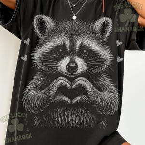 Peut inclure: T-shirt noir avec une illustration en niveaux de gris d'un raton laveur formant un c&oelig;ur avec ses mains. Le motif comprend de petits c&oelig;urs blancs et le texte "THE LUCKY SHAMROCK".