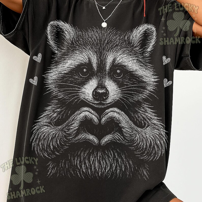 Peut inclure: T-shirt noir avec une illustration en niveaux de gris d'un raton laveur formant un c&oelig;ur avec ses mains. Le motif comprend de petits c&oelig;urs blancs et le texte "THE LUCKY SHAMROCK".